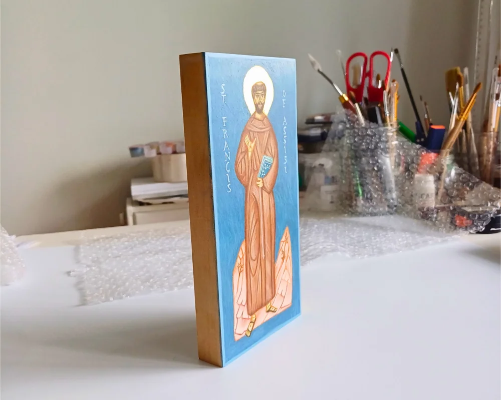 Hand-painted St. Francis of Assisi icon on wood, featuring the full-body saint with stigmata, nature background, and Bible, photographed in a professional iconography studio with brushes and pigments.
