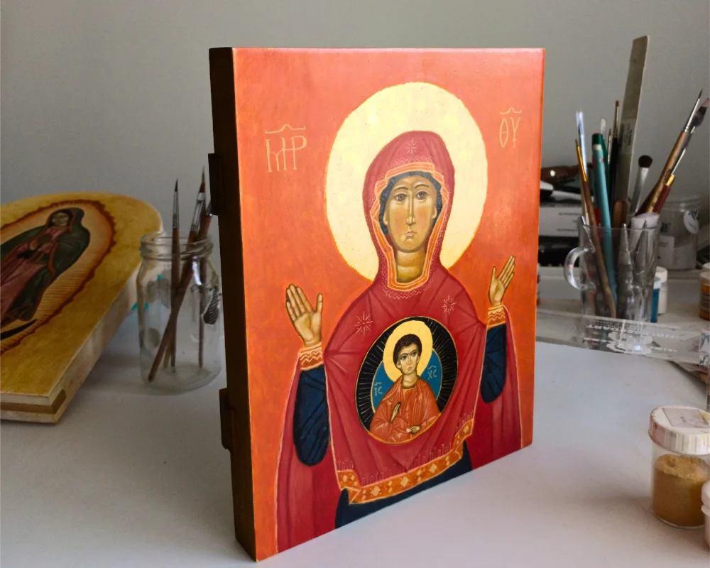 A handmade Mother of God Orant icon with 24k gold leaf on a wooden panel, displayed in a professional icon painting studio with brushes in the background.