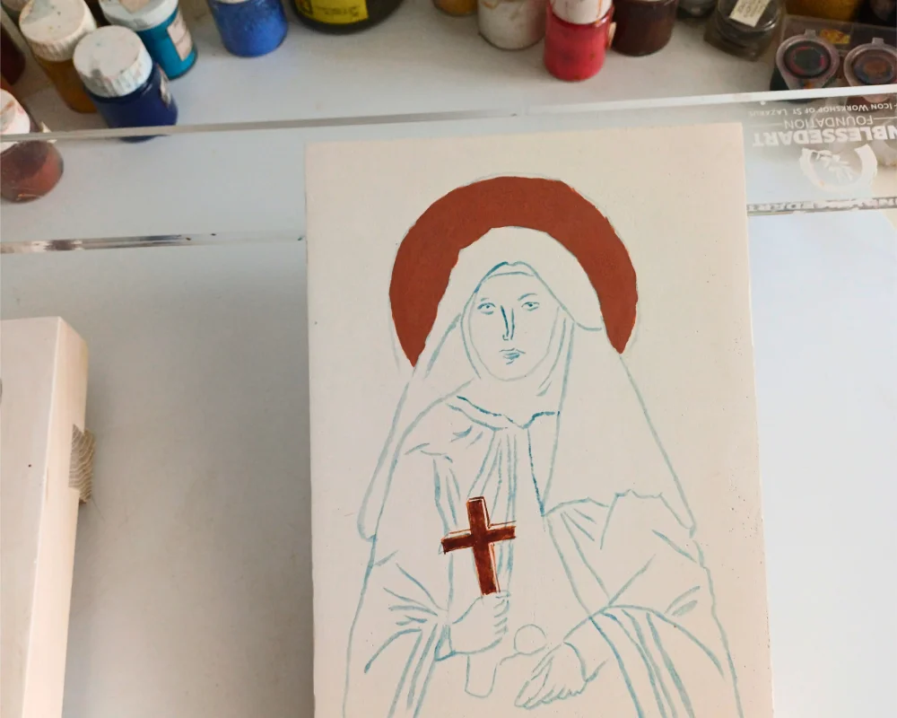 First stage of writing a St Therese of Lisieux hand-painted icon with a red clay bolus for gold leafing and a small crucifix held in hand.